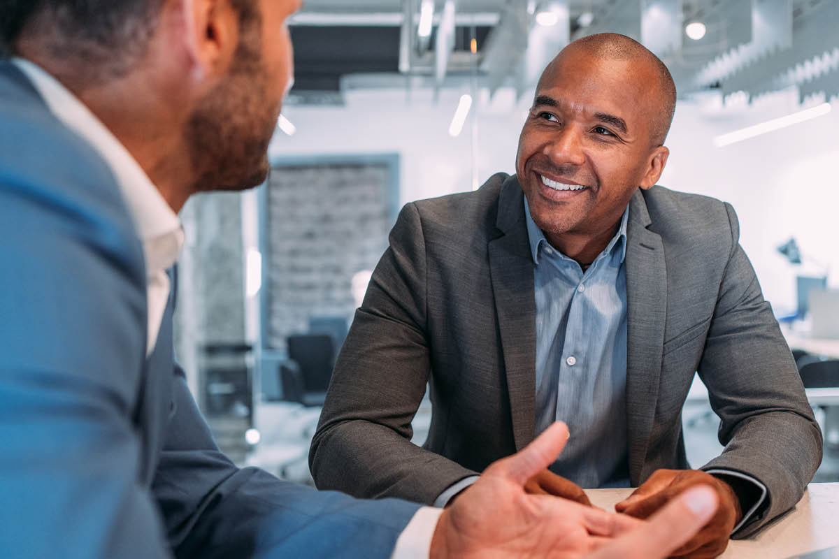 The image captures a cheerful business conversation between two individuals. The man in focus is smiling broadly and has a confident, engaging demeanor. 