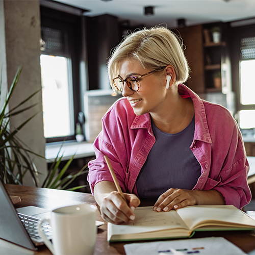 Young woman with glasses looking at laptop and taking notes. She is studying professional development courses on-demand at home.
