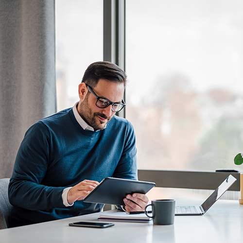 Young businessman using digital tablet while working on laptop in business office. Male professional with wireless computer at desk. He is sitting by window in office.