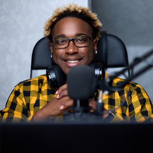 A person sitting on a chair in a yellow and black shirt. They have headphones resting on their back and a microphone in front of them. 
