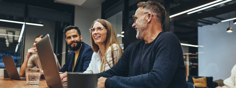 An image of people seated in a room smiling while on their laptops.
